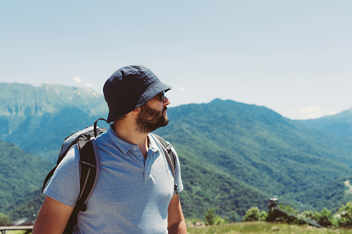 millenial bearded tourist man wearing bucket hat, with backpack trekking in mountains