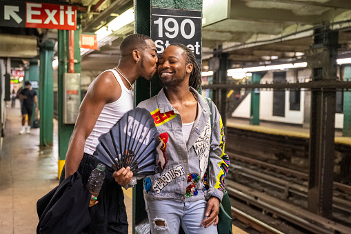 Gay New Yorkers in 20s and 40s standing underground at 190th Street station, sharing affection and smiling, summertime.