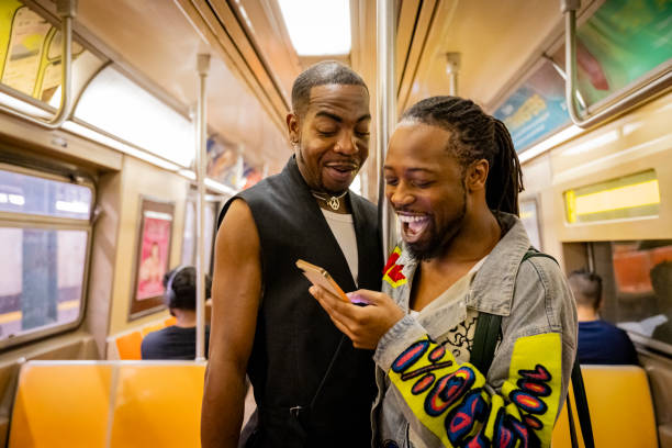 Gay New Yorkers in 20s and 40s, dressed for summer leisure, looking at smart phone and laughing with expressions of surprise.