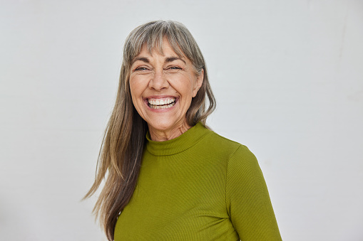 Portrait of a mature woman wearing a green shirt laughing while standing in front of a light gray background