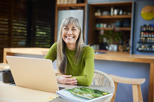 Portrait of a mature woman laughing and working on a laptop while sitting at a dining table in her kitchen at home