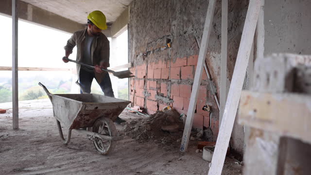 Latin American construction worker picking up materials with a shovel into a wheelbarrow at a building site