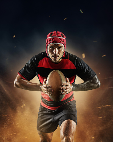 Young man, professional rugby player in uniform standing with rugby ball over dark stadium with flashlight and mist. Concept of professional sport, competition, motivation, game, championship