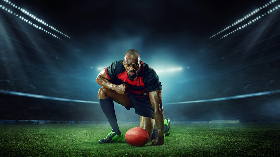 Senior, concentrated, young, African-American man, rugby player sitting on filed over dark empty stadium. Winner. Concept of professional sport, competition, motivation, game, championship
