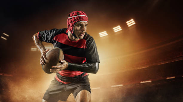 istock Concentrated young man, rugby player in uniform with ball standing on dark empty field with flashlights and mist 1796577963