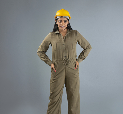 Portrait of confident female architect dressed in coveralls and hardhat standing with hands in pockets against gray background