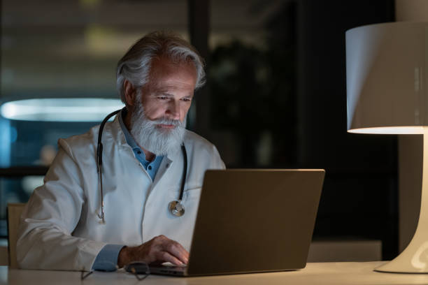 lekarz nocnej zmiany bada przypadek na laptopie w zaciemnionym biurze - doctors office examination table medical exam clinic zdjęcia i obrazy z banku zdjęć