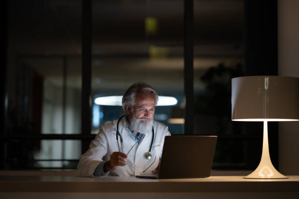 um médico do turno da noite examina um caso em um laptop em um consultório escuro - doctors office examination table medical exam clinic - fotografias e filmes do acervo