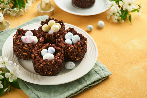 istock Easter chocolate nest cake with mini chocolate candy eggs with blossoming cherry or apple flowers on yellow background table. Creative recipe for Easter table with holiday decorations. Top view. 1796623452