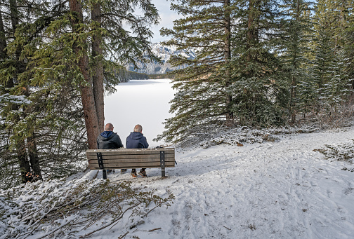 Banff National Park, Alberta, Canada – November 12, 2023:  Two men sit on a bench overlooking Johnson Lake