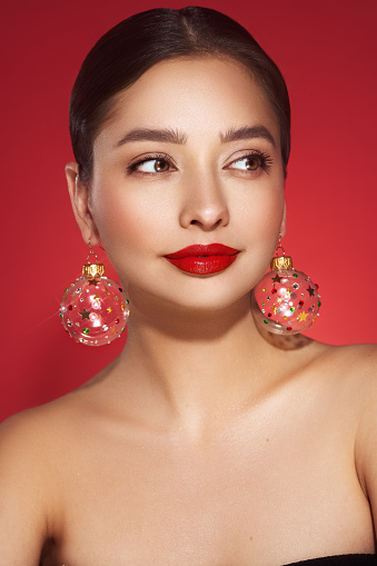 Portrait of beautiful young woman on red background, red lipstick and festive makeup, Christmas toy earrings, shiny glowing skin, Winter holidays concept