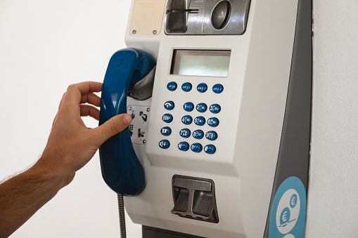 Old public telephone for making voice calls using coins or prepaid card stuck on a white wall. Person's hand holding handset to make a call.