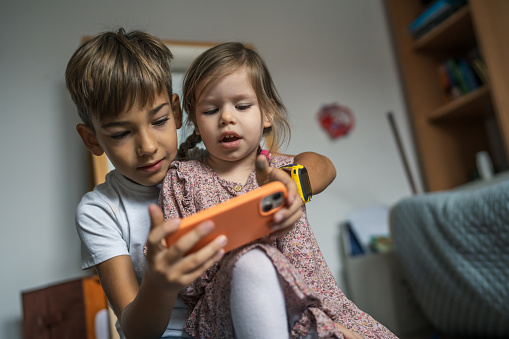 Siblings brother boy hold smartphone with girl sister children use mobile phone smartphone at home in room watch video make a call or play online games leisure family concept real people copy space