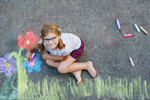 Cute little girl and flowers painted with colorful chalks on asphalt. Happy preschool child having fun with painting chalk picture. Creative leisure for children, drawing and painting