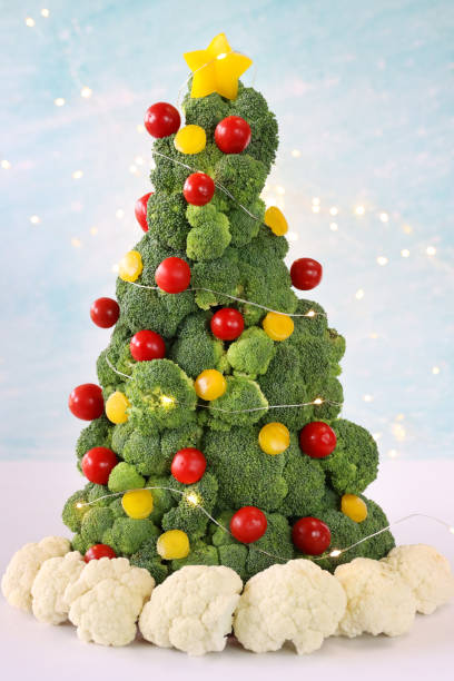 istock Close-up image of broccoli Christmas tree buffet centrepiece with cauliflower snow base, yellow bell pepper and red cherry tomato baubles, illuminated fairy lights, pale blue background, focus on foreground 1796785797