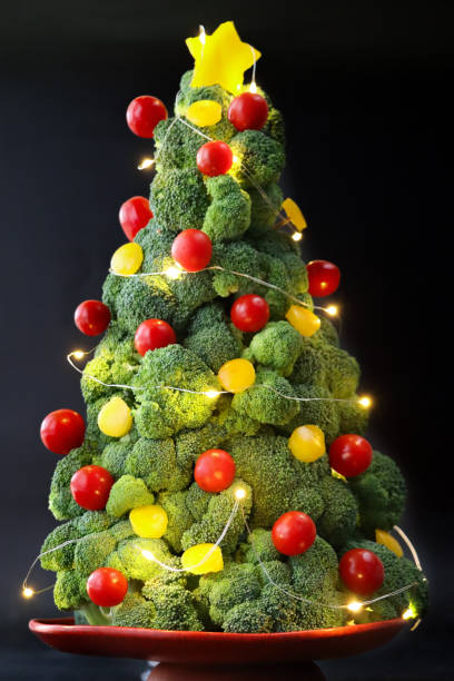 istock Close-up image of broccoli Christmas tree buffet centrepiece on stand, yellow bell pepper and red cherry tomato baubles, illuminated fairy lights, black background, focus on foreground 1796786334