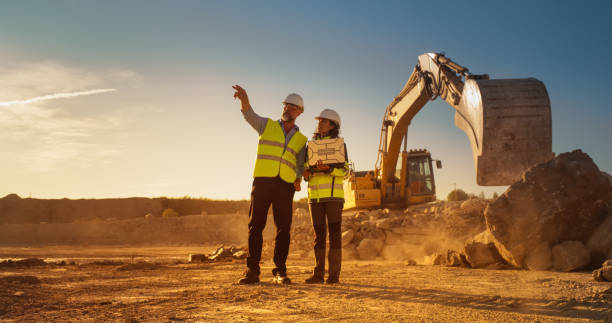 istock Caucasian Male Civil Engineer And Hispanic Female Architect Walking On Construction Site With Laptop Computer And Talking About New Real Estate Project. Machinery Working On Background. Golden Hour. 1796830561