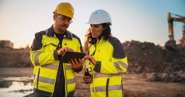 Caucasian Male Civil Engineer Talking To Hispanic Female Inspector And Using Tablet Computer On Construction Site of a New Building. Real Estate Developers Discussing Business, Excavators Working.