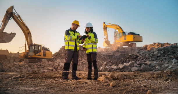 kaukasischer männlicher bauingenieur spricht mit hispanischer inspektorin und benutzt tablet auf der baustelle eines neuen apartmentkomplexes. immobilienentwickler diskutieren über geschäfte, bagger arbeiten. - schwerindustrie stock-fotos und bilder