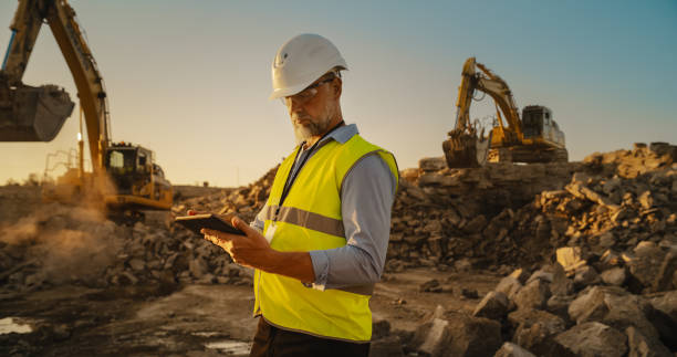 istock Caucasian Male Civil Engineer Wearing Protective Goggles And Using Tablet On Construction Site On A Sunny Day. Man Inspecting Building Progress. Excavator Loading Materials Into Big Industrial Truck. 1796833756