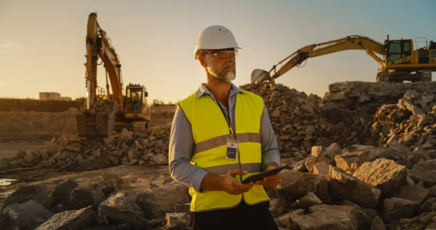 istock Caucasian Male Civil Engineer Wearing Protective Goggles And Using Tablet On Construction Site On A Sunny Day. Man Inspecting Building Progress. Excavator Loading Materials Into Industrial Truck. 1796833767