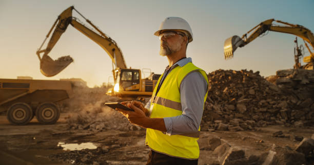 istock Caucasian Male Civil Engineer Wearing Protective Goggles And Using Tablet On Construction Site On Sunny Day. Man Inspecting Building Progress. Excavator Loading Materials Into Big Industrial Truck. 1796834278