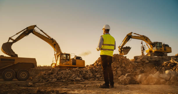 caucasian male urban planner wearing protective goggles and using tablet on construction site on a sunny day. man inspecting building progress. excavator loading materials into industrial truck - i̇nşaat sanayisi stok fotoğraflar ve resimler