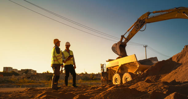 istock Caucasian Real Estate Investor With Tablet And Project Manager Talking On Construction Site Of Apartment Building. Colleagues Wearing Hard Hats And Watching Heavy Machinery Working. Sunny Day 1796838697