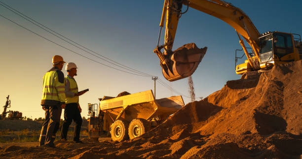 istock Caucasian Real Estate Investor With Tablet Computer And Project Manager Talking On Construction Site Of Apartment Building. Colleagues Wearing Hard Hats And Watching Heavy Machinery Working. Sunny Day 1796838726