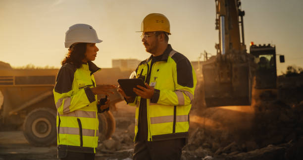 plan cinématographique de l’heure dorée du chantier de construction : un ingénieur civil caucasien et une urbaniste hispanique parlent, à l’aide d’une tablette. camions, excavatrices, chargeuses travaillant à la construction d’un nouveau quar - vérifier lheure photos et images de collection