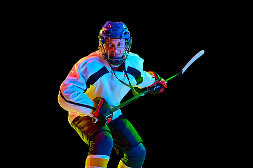 Concentrated, motivated young girl, hockey player standing in uniform with stick against black studio background in neon light. Concept of professional sport, competition, game, action, hobby