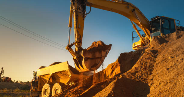istock Construction Site On Sunny Evening: Industrial Excavator Loading Sand Into A Truck. The Process Of Building New Apartment Block. Workers Operating Heavy Machinery To Complete Real Estate Project. 1796843561
