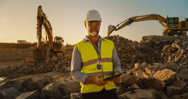 istock Successful Bearded Civil Engineer Wearing Protective Goggles And Looking At Camera On Construction Site on a Sunny Day While Heavy Machinery Working. Man Holding Tablet, Wearing Hard Hat, Safety Vest. 1796847318