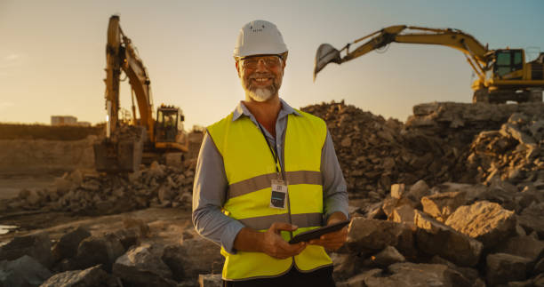 istock Successful Bearded Civil Engineer Wearing Protective Goggles And Smiling At Camera On Construction Site on a Sunny Day While Heavy Machinery Working. Man Holding Tablet, Wearing Hard Hat, Safety Vest. 1796847850