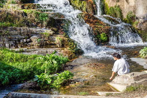 a girl in a white T-shirt sits next to a small waterfall in Tbilisi