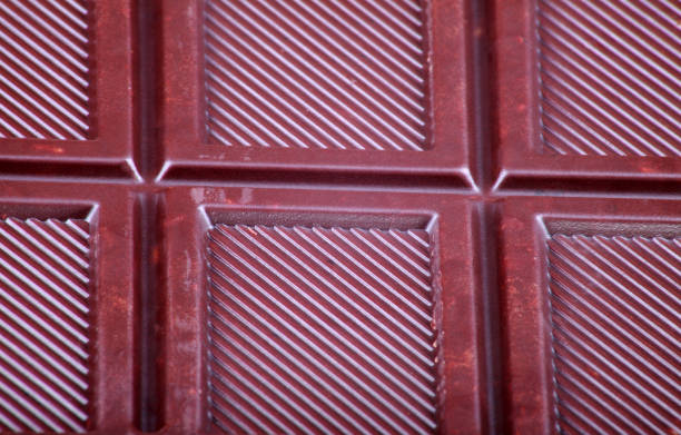 A milk chocolate bar with a broken piece placed on wavy desert sand, creating a striking visual contrast between the creamy cocoa texture and the smooth, flowing dunes. The image captures the warmth and richness of chocolate in a conceptual gourmet food photography setting, emphasizing indulgence, natural beauty, and the interplay of textures.