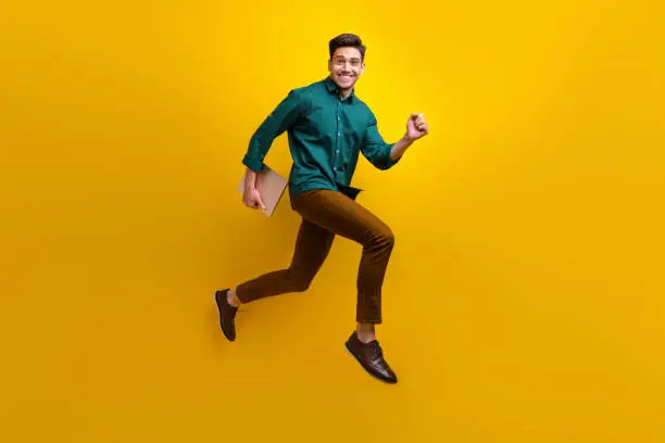 Photo of Photo of cheerful nice man wear stylish clothes run hold netbook macbook isolated on yellow color background