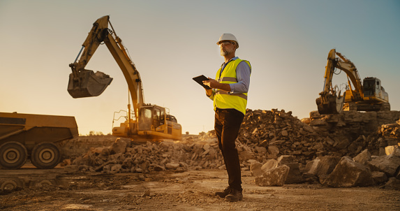 Caucasian Male Real Estate Investor Wearing Protective Goggles And Using Tablet On Construction Site On Sunny Day. Man Inspecting Building Progress. Excavator Loading Materials Into Industrial Truck