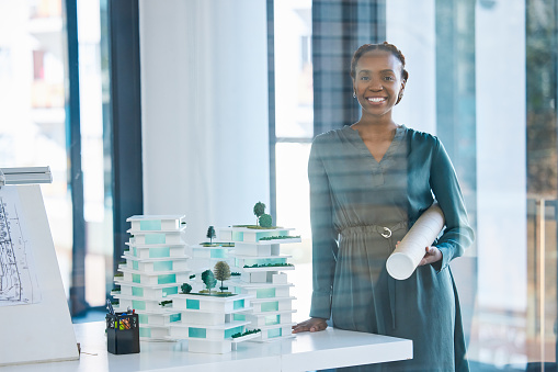 Architect, engineer and designer holding a blueprint with a positive mindset, mission and vision in an office. Portrait of a black woman standing next to 3D representation of proposed building design