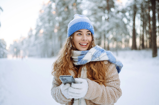 A young woman in a snowy park with a phone in her hands communicates via video and takes a selfie. Beautiful female tourist with phone having fun outdoors. Travel concept.