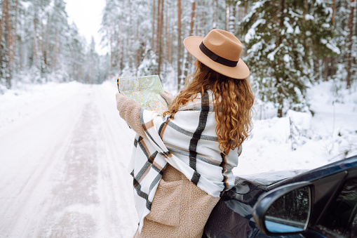 Happy woman in a hat with a map near the car on a snowy winter road in the forest. A traveler is looking for directions using a map on a winter day. Car travel concept.
