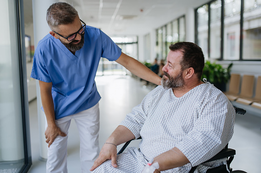 Close up of supportive doctor talking with worried overweight patient in wheelchair. Illnesses and diseases in middle-aged men's health. Compassionate physician supporting stressed patient. Concept of health risks of obesity.