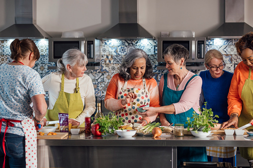 A group of mature and senior women enjoying a staycation and taking part in a cooking class in the accommodation they're staying at in Amble, North East England. The main focus is one woman about to take an ingredient out a pot that her friend is holding for her.

Videos also available for this scenario.