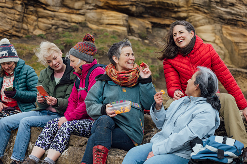 A group of mature and senior women enjoying a day out in Howick, North East England. They are taking a break from exploring, sitting on a rock formation, enjoying a fruit snack and some of the other women are looking at mobile phones.\n\nVideos also available for this scenario.