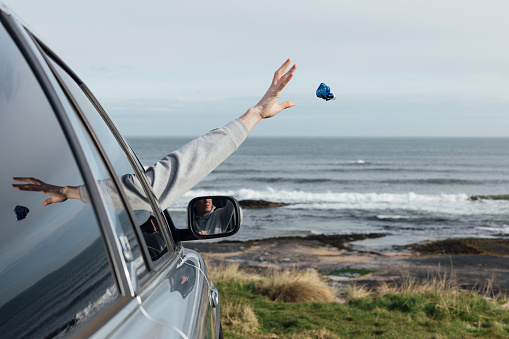 An unrecognisable person throwing litter out their car window as they sit next to the coastline in Bamburgh, North East England. Their arm is raised and reaching straight out the window after letting the rubbish packet out of their hand with the sea and beach in the background of the shot.

Videos also available for this scenario.