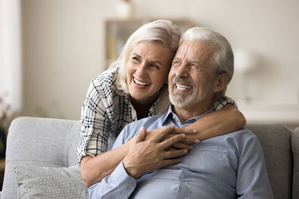 Dreamy elderly aged couple in love hugging at home, laughing Dreamy elderly aged couple in love hugging at home, laughing, looking away in happy thoughts, thinking on good retirement plans, discussing vacation, enjoying marriage, relationships teeth bonding stock pictures, royalty-free photos & images