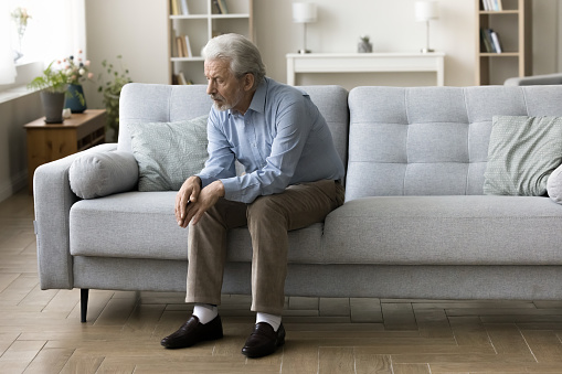 Sad lonely old grey haired man sitting alone on home sofa, looking down in bad thought, suffering from depression, apathy, coping with loss, bad news about healthcare, family problems