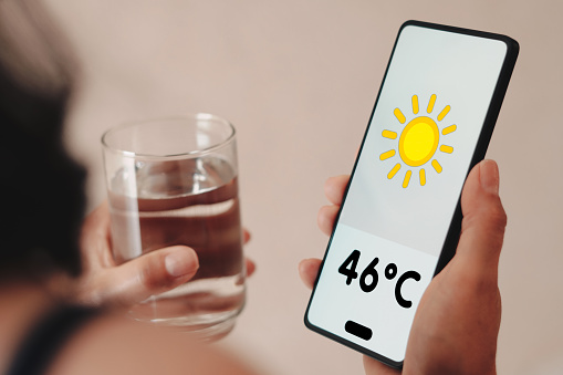 A woman holds a glass of water, the thermometer showing 46ºC of heat is displayed on the screen of a smartphone.