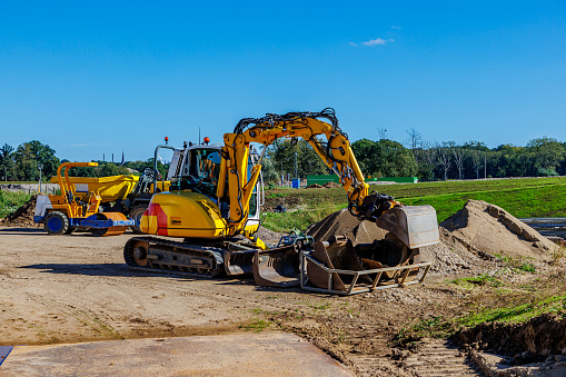 Yellow crawler excavator on working ground in construction of rural road, working machinery, agricultural land and trees against blue sky in background, sunny day in Meers, Elsloo, Netherlands