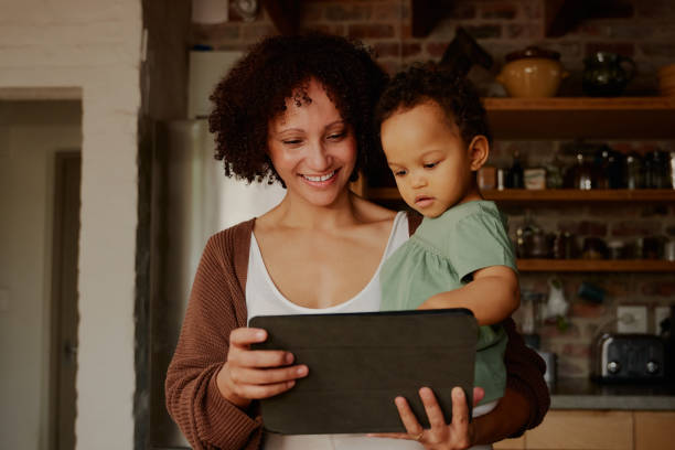 mère biraciale heureuse portant sa fille tout en utilisant une tablette numérique dans la cuisine à la maison - placard holding celebration women photos et images de collection